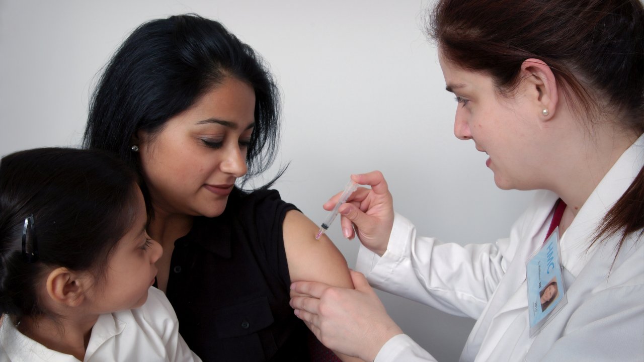 Foto de mulher aplicando vacina do HPV em outra mulher, com criança no colo