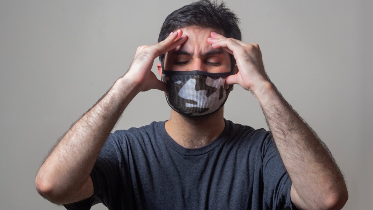 Foto de homem com máscara e mãos na cabeça, simbolizando enxaqueca