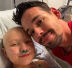 Mulher com cabelo raspado, usando cânula respiratória em casa de hospital. Ao seu lado, homem de camiseta vermelha.