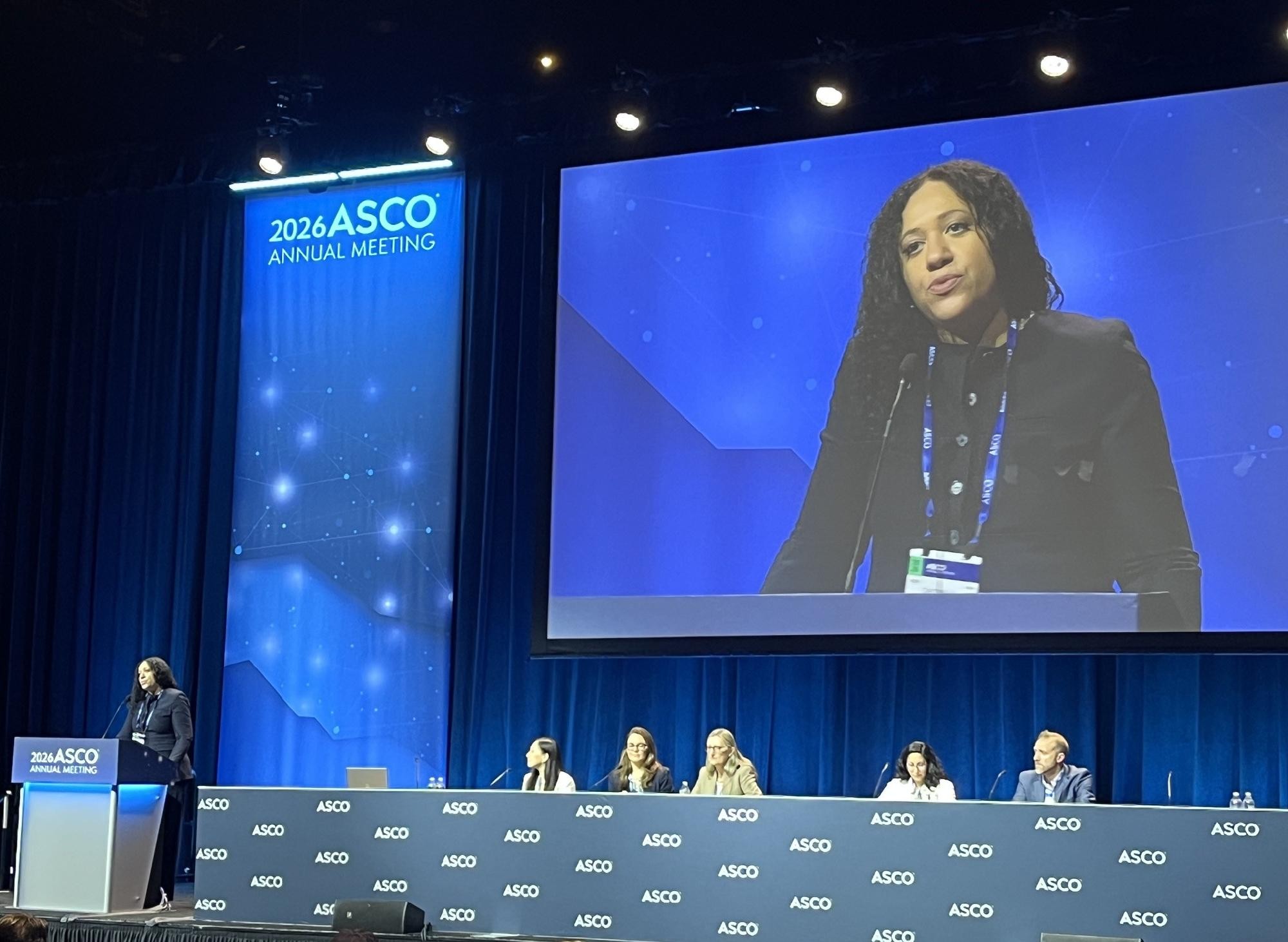 Mulher negra de cabelos cacheados e blazer preto discursa em pódio num palco azul, com a imagem dela projetada em telão gigante ao fundo. Abaixo, cinco pessoas sentadas em mesa com o logo ASCO repetido