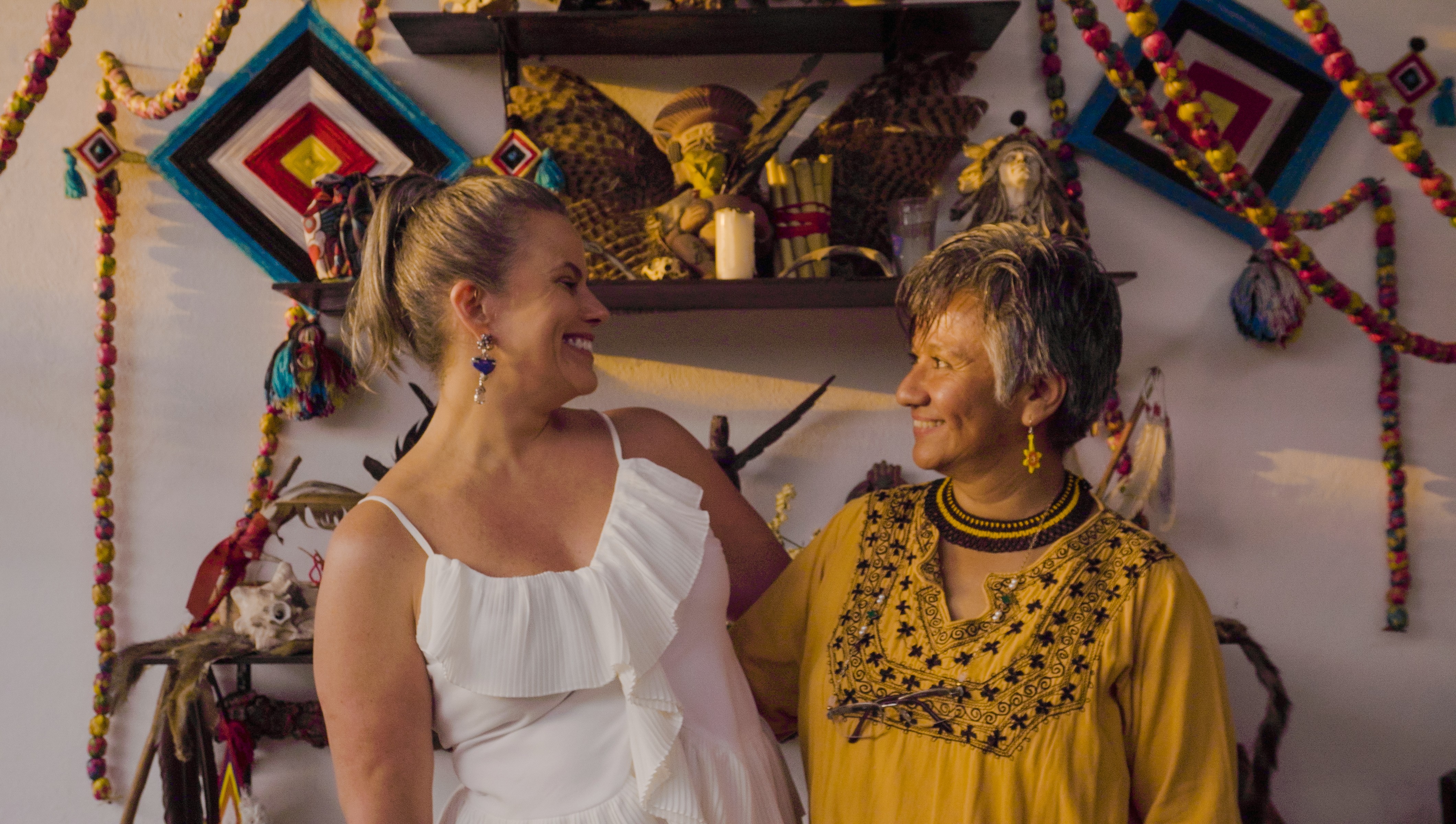 Duas mulheres sorrindo, uma loira de vestido branco e outra de cabelos grisalhos com blusa amarela bordada, em ambiente decorado com artesanato indígena