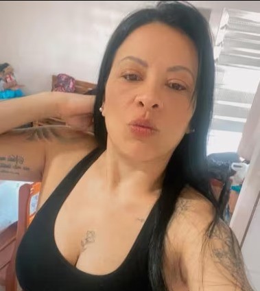 Mulher de cabelos pretos longos, com tatuagens nos braços e no peito, vestindo top preto, fazendo bico para a câmera em um ambiente interno