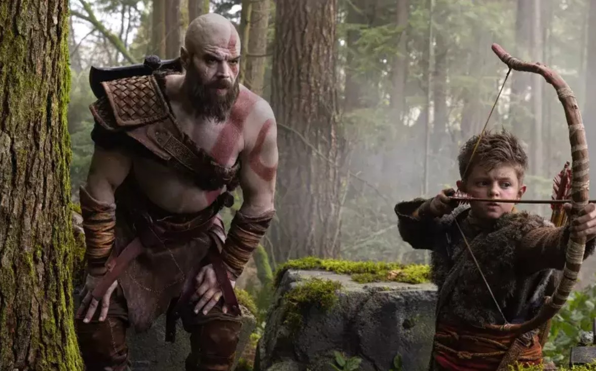 Kratos, um homem careca e barbudo com marcas vermelhas no corpo, observa Atreus, um menino loiro com arco e flecha, em uma floresta densa e úmida