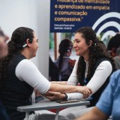Duas mulheres com cabelos cacheados e fones de ouvido conversam sorrindo, sentadas em carteiras escolares, com as mãos entrelaçadas sobre a mesa. Ao fundo, um banner azul com texto em português sobre empatia e comunicação compassiva. Elas estão fazendo uma simulação do projeto Simfonik.