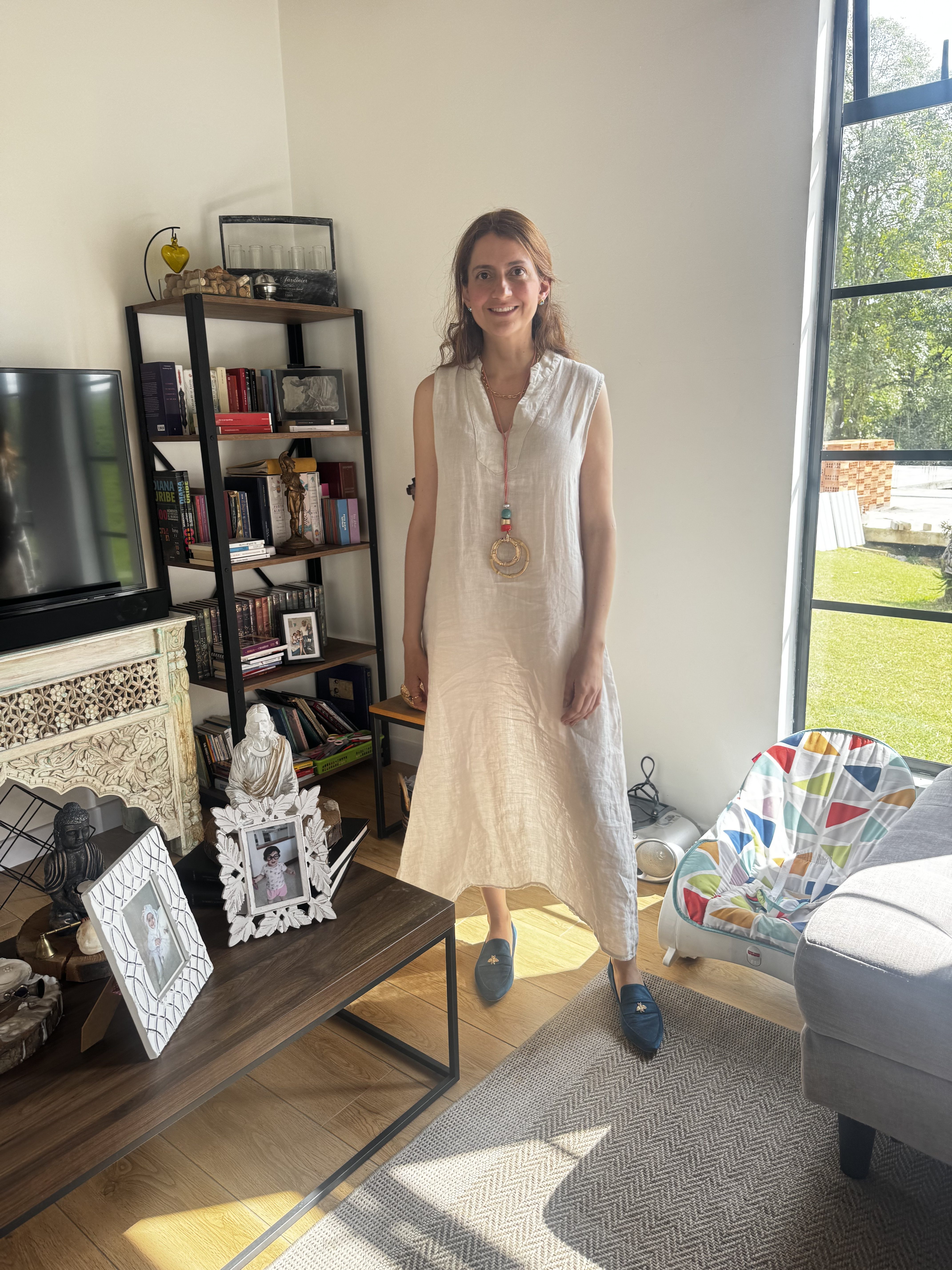 Mulher sorridente, de cabelos castanhos e vestido bege claro, em pé na sala de estar. Ela usa um colar com pingente circular e miçangas coloridas. Ao fundo, uma estante com livros e objetos decorativos, uma TV e uma janela grande com vista para o jardim. No chão, um tapete cinza e um berço colorido
