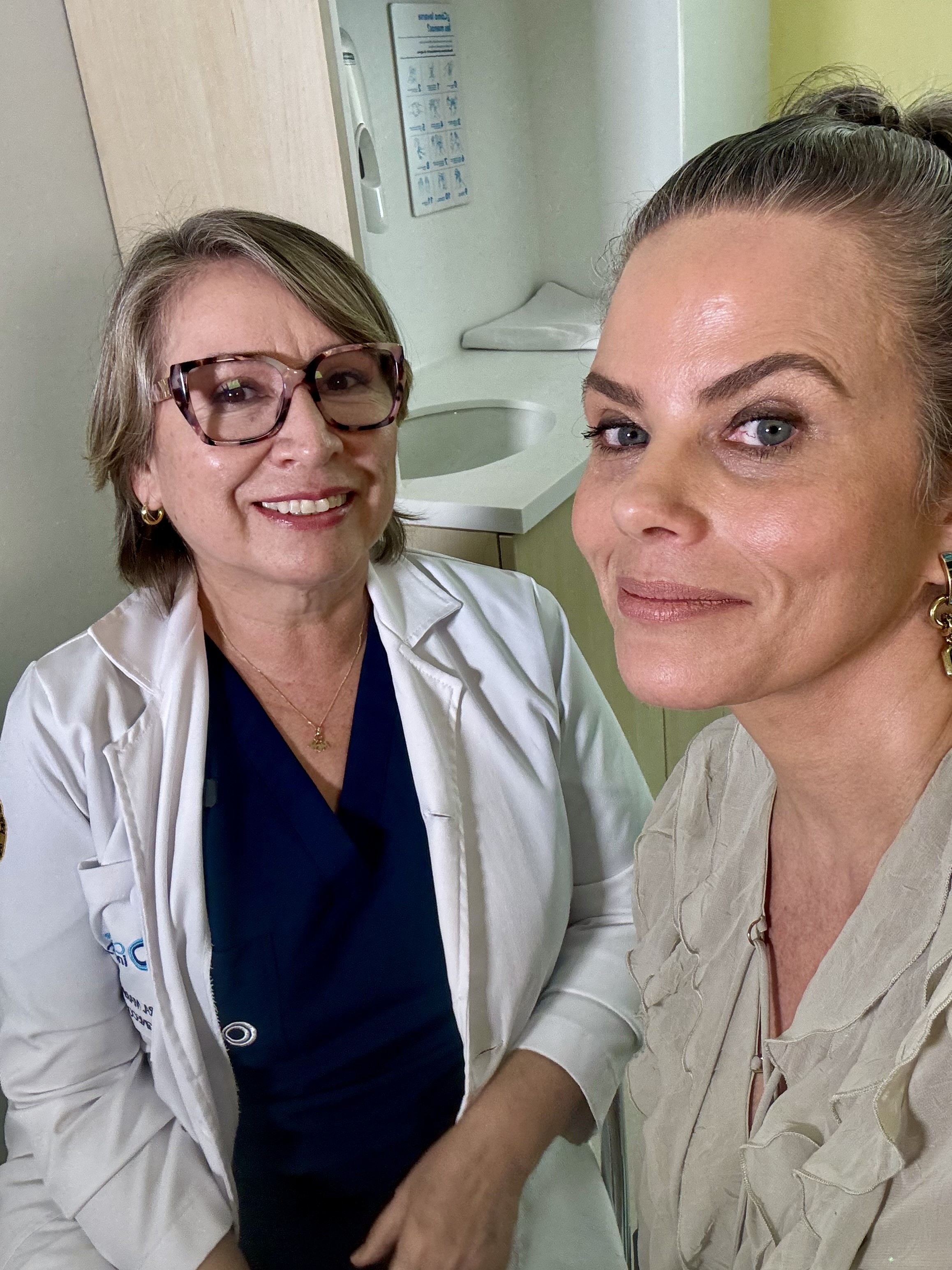 Duas mulheres sorrindo para a câmera em um ambiente médico. À esquerda, uma médica de jaleco branco e óculos, com um colar dourado. À direita, uma mulher com blusa bege e brincos dourados, cabelo preso. Ao fundo, uma pia e um dispenser de papel.