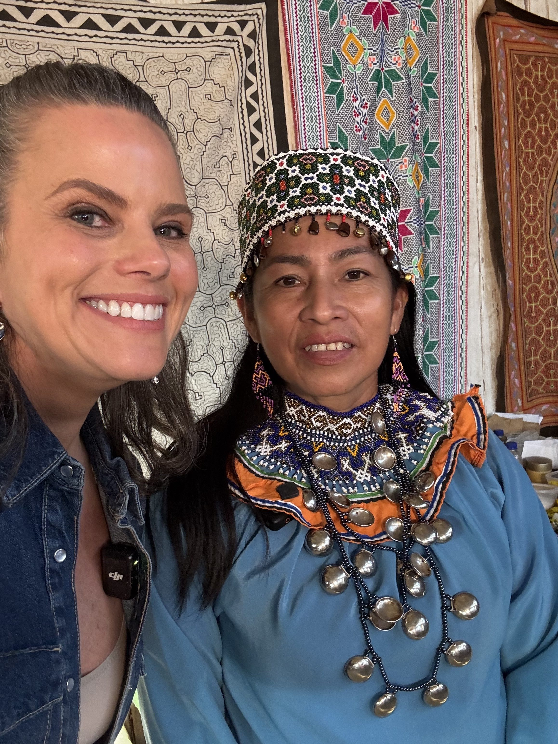 Duas mulheres sorrindo para a câmera, uma com chapéu bordado e roupa azul com colar de moedas, a outra com jaqueta jeans, sobre tapetes estampados