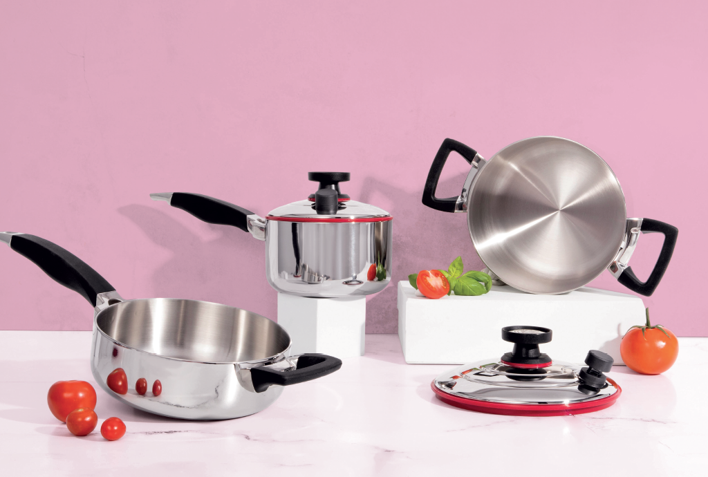 Conjunto de panelas de aço inox com detalhes vermelhos e cabos pretos, dispostas sobre uma superfície de mármore branco com fundo rosa. Há tomates e folhas de manjericão espalhados
