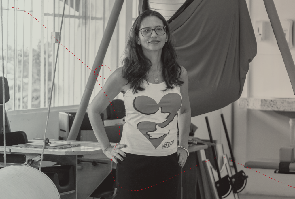 Mulher de óculos e cabelos longos, sorrindo, com as mãos na cintura, vestindo regata branca com estampa de coração e saia escura, em ambiente interno com equipamentos de ginástica e uma rede de tecido pendurada