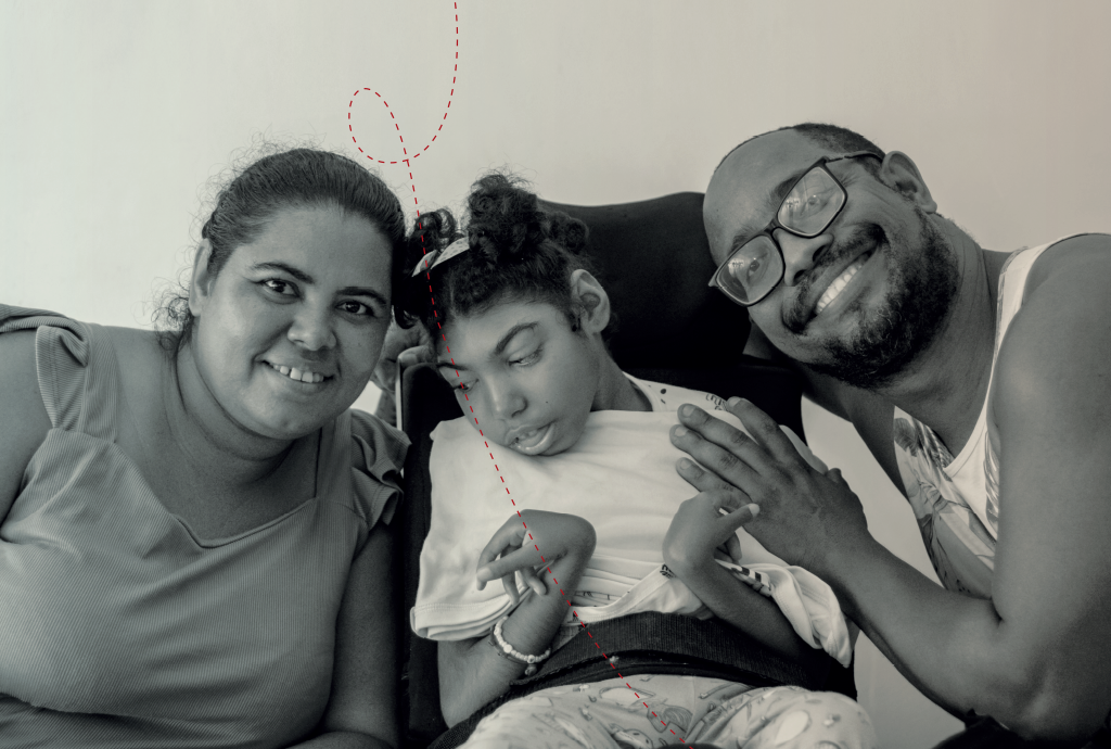 Família sorridente em preto e branco: mulher, criança com deficiência e homem. A mulher e o homem abraçam a criança, que está sentada em uma cadeira adaptada. O homem usa óculos e a mulher tem cabelo preso. Todos olham para a câmera com expressões felizes