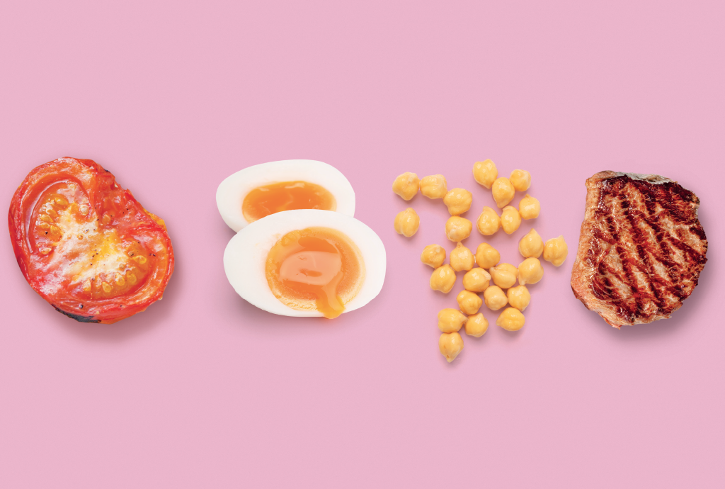 Metade de um tomate assado, dois ovos cozidos com gema mole, grãos de grão-de-bico e um bife grelhado, dispostos em linha sobre um fundo rosa