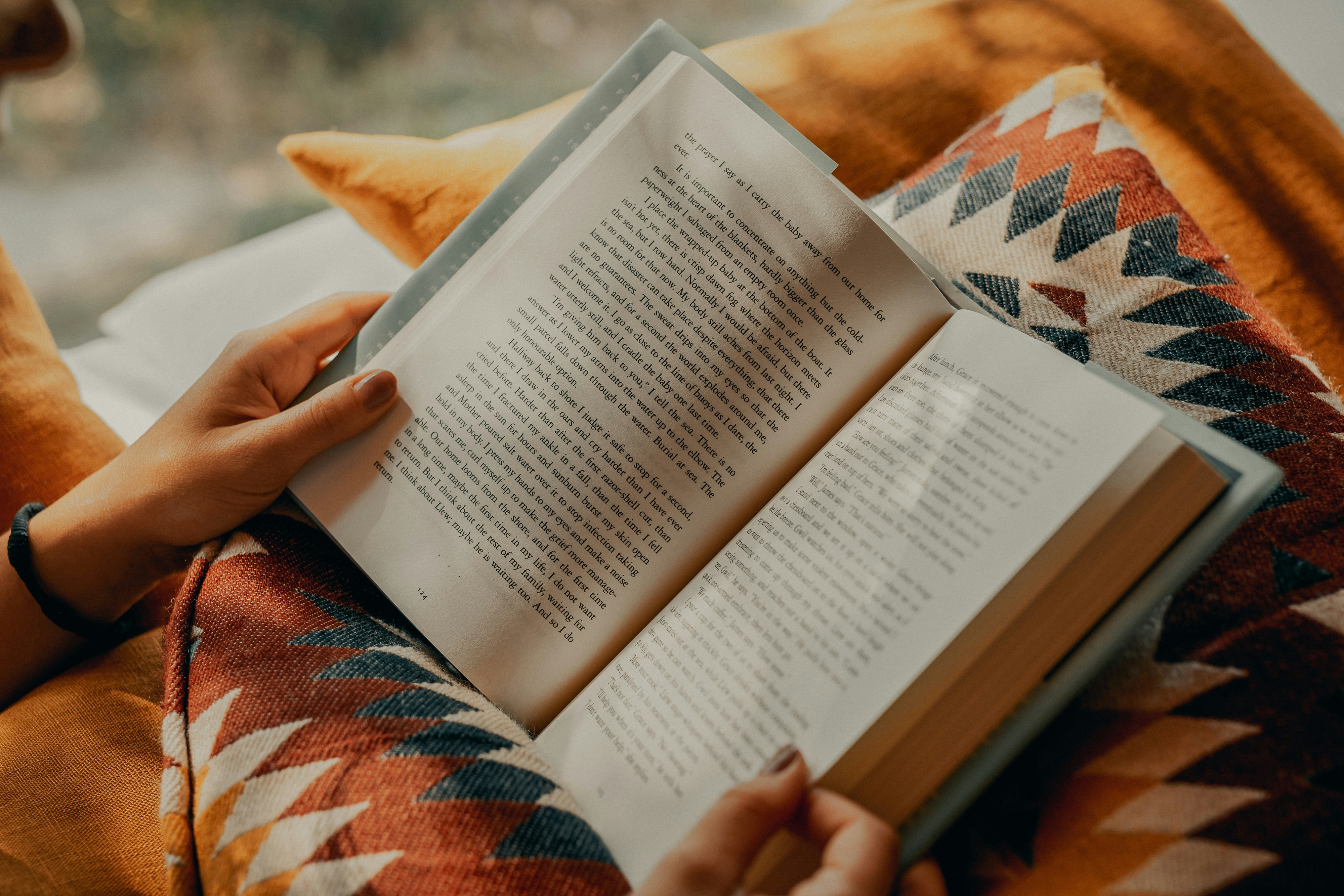 Mãos de uma pessoa segurando um livro aberto, com páginas claras e texto em inglês, sobre um cobertor ou almofada com estampa geométrica em tons de laranja, azul e branco, e um travesseiro amarelo ao fundo