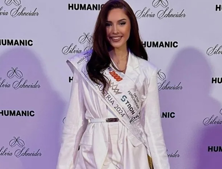 Mulher jovem de cabelos castanhos longos, sorrindo, vestindo um blazer branco e uma faixa branca com Miss Austria 2024 e logotipos, em frente a uma parede com os nomes Humanic e Silvia Schneider repetidos