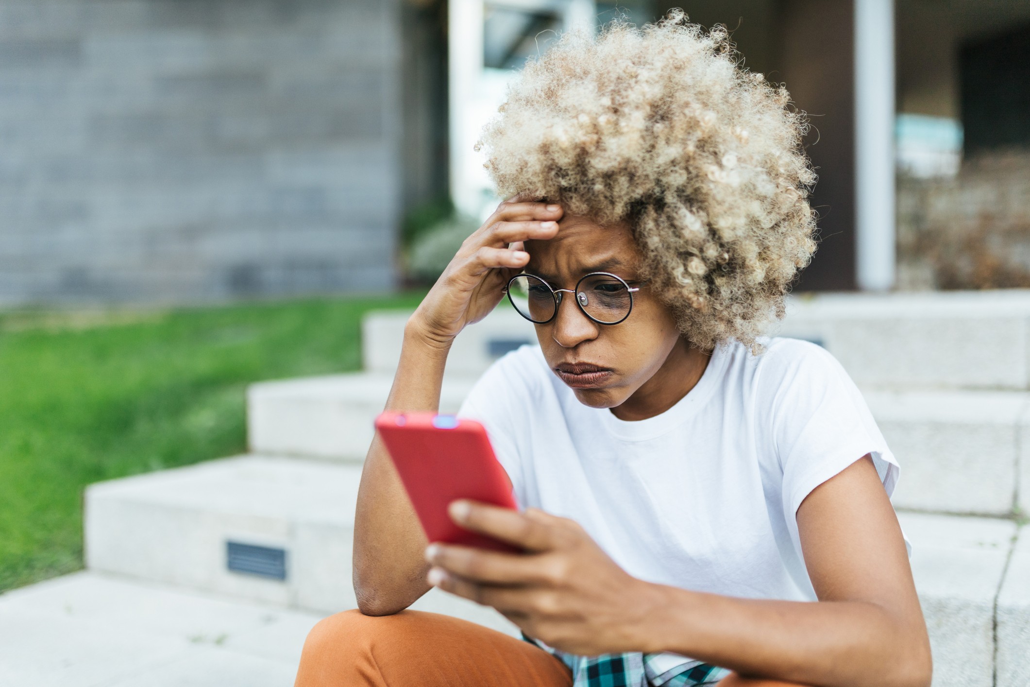 Mulher negra com cabelo crespo loiro, óculos redondos e camiseta branca, sentada em degraus de concreto, olhando para um celular vermelho com expressão preocupada, mão na testa