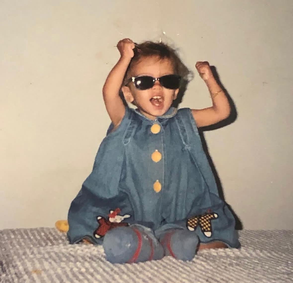 Criança pequena de pele morena, cabelo escuro e óculos de sol pretos, vestindo um macacão jeans com botões amarelos e aplicações de bichinhos, sentada em uma cama com lençol xadrez cinza e branco, com os braços levantados e a boca aberta em um grito de alegria