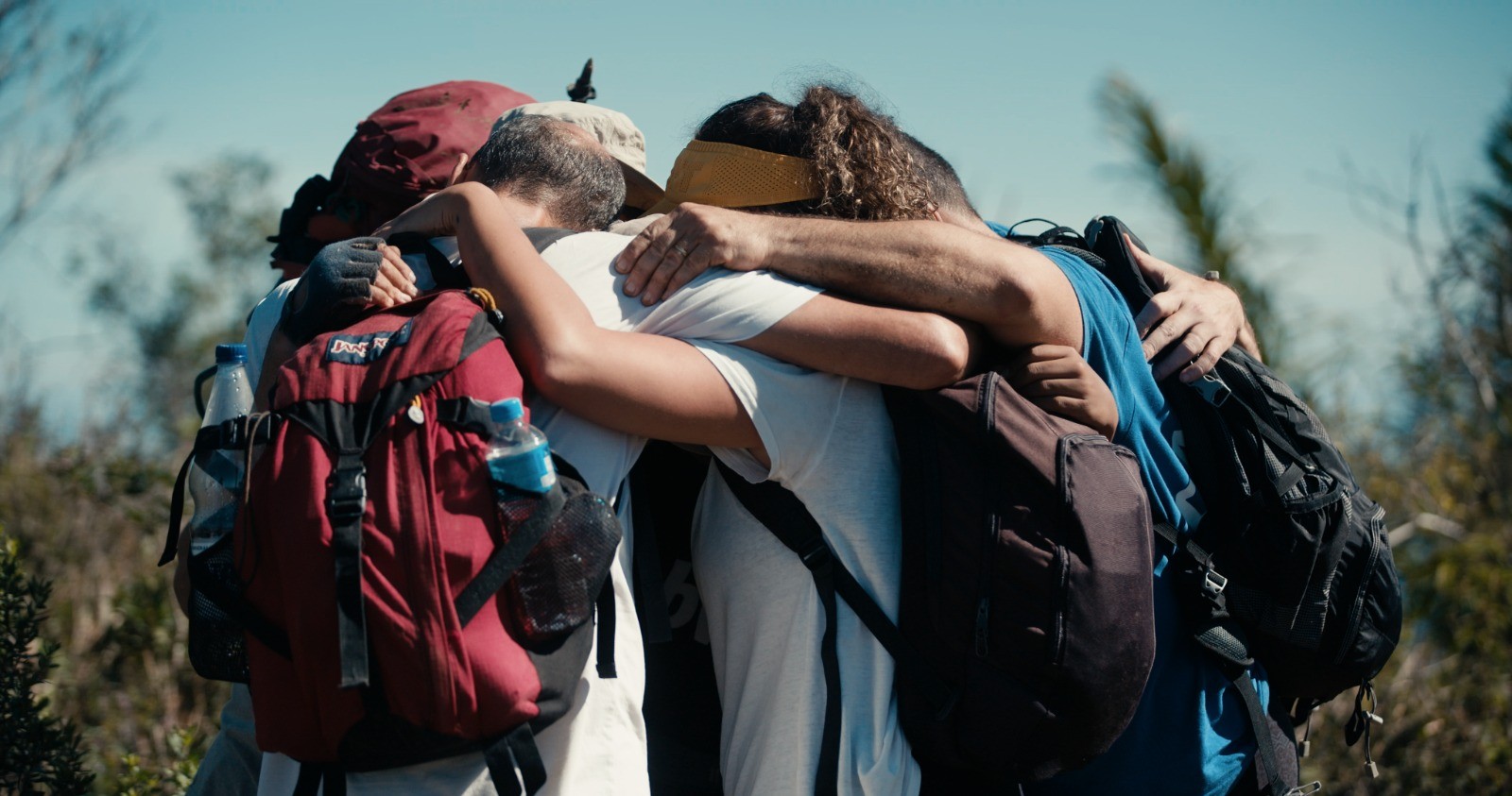 Quatro pessoas, com mochilas de trilha e garrafas de água, abraçam-se em grupo, de costas para a câmera, sob céu azul claro e vegetação embaçada ao fundo