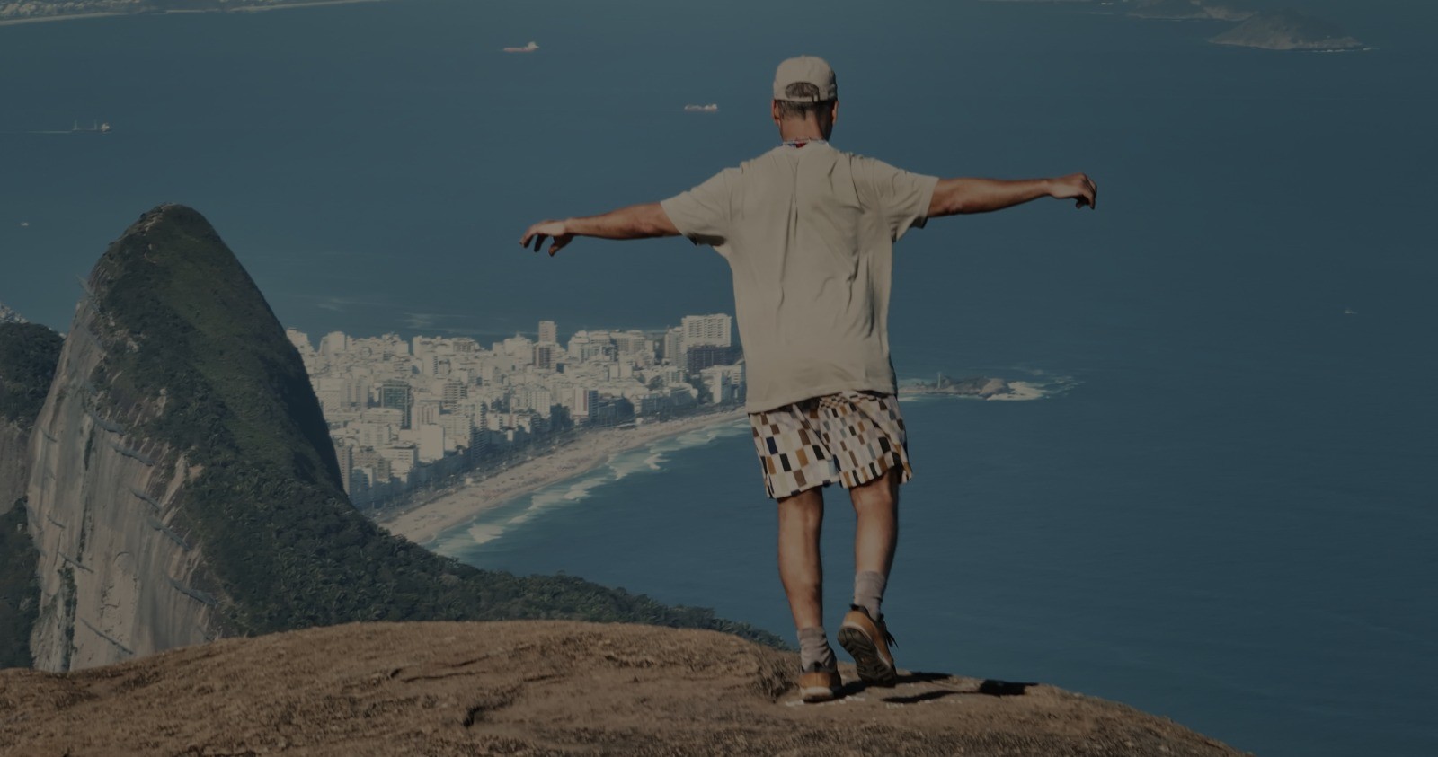Homem de boné e bermuda colorida, de costas, braços abertos, equilibrando-se na beira de um penhasco rochoso. Ao fundo, a cidade do Rio de Janeiro com prédios, praia e o mar azul com barcos.